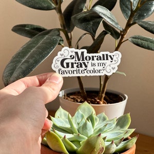 May include: A white sticker with black text that reads "Morally Gray is my favorite color." The sticker has a decorative border and small hearts around the text.