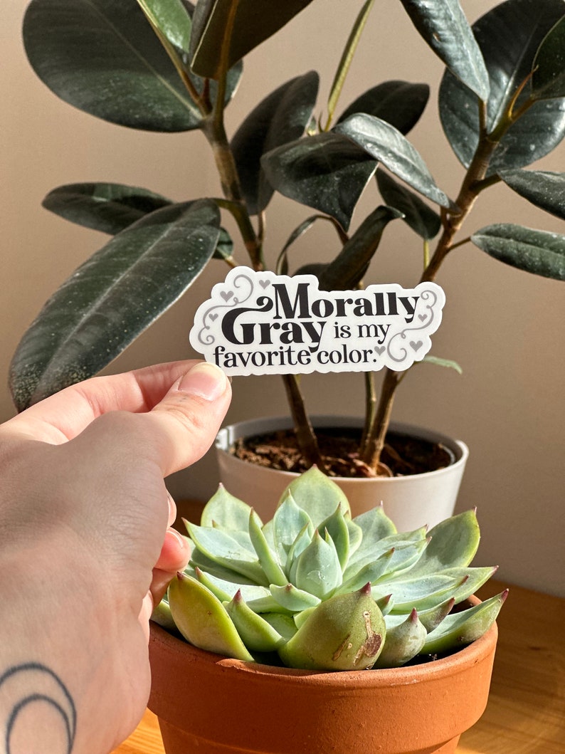 May include: A white sticker with black text that reads "Morally Gray is my favorite color." The sticker has a decorative border and small hearts around the text.