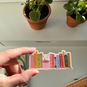 May include: A sticker of a stack of books with titles like "Soul Mates", "Love Triangle", and "Fake Dating". The books are stacked on top of a small pot with a white flower.