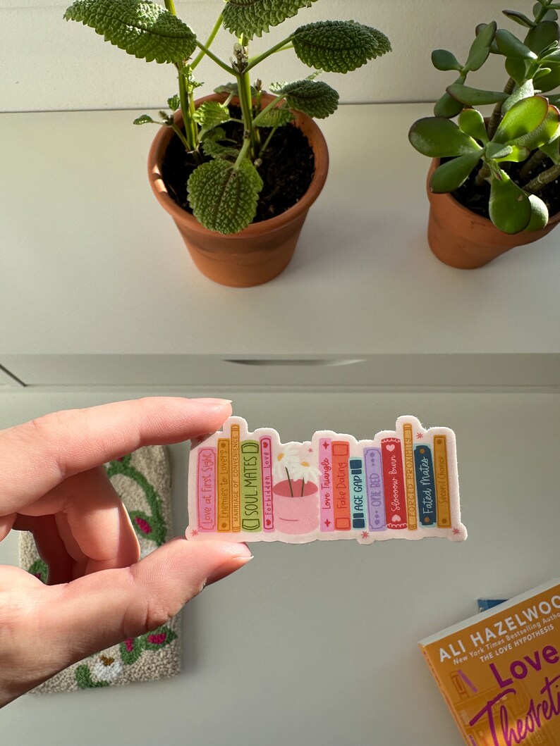 May include: A sticker of a stack of books with titles like "Soul Mates", "Love Triangle", and "Fake Dating". The books are stacked on top of a small pot with a white flower.