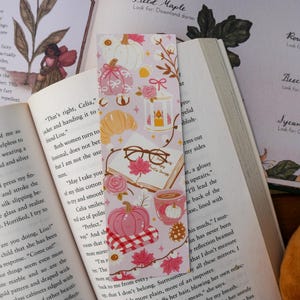 May include: A pink bookmark with autumn-themed illustrations, including pumpkins, maple leaves, and a croissant. It features a cup of coffee, glasses, and the text "Pumpkin Spice Things".