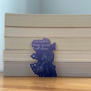 May include: A purple sticker with a line drawing of a person holding a book. The text on the sticker reads "we read to know we are not alone".