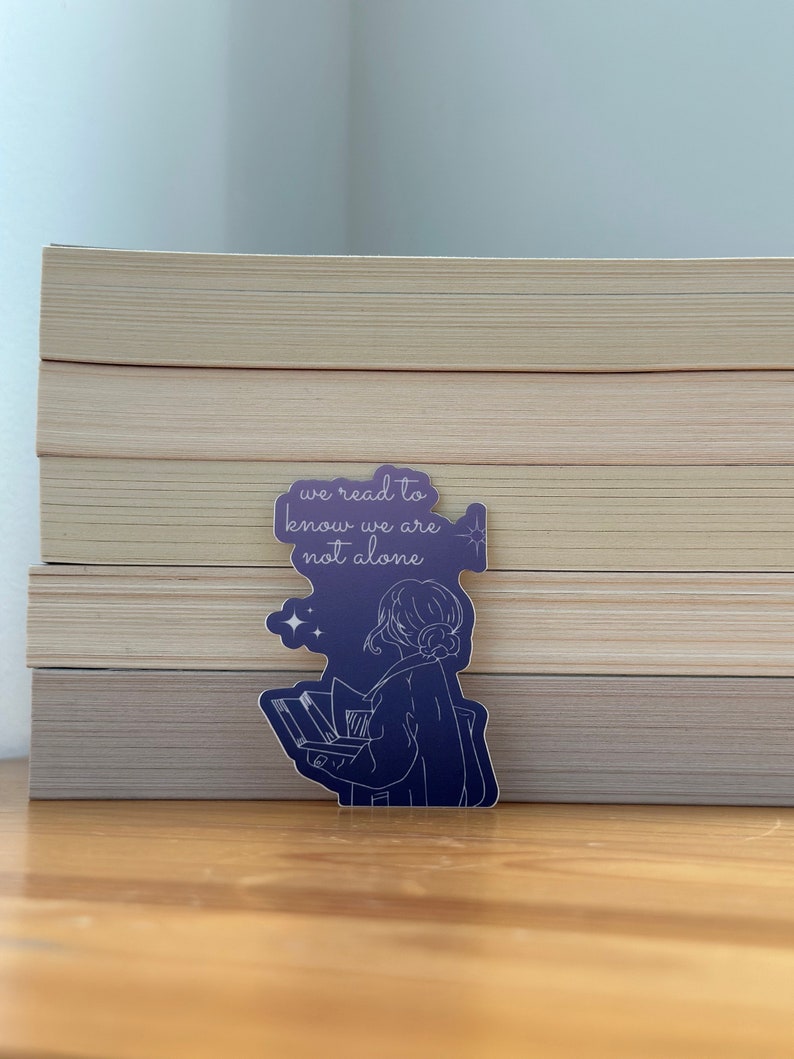 May include: A purple sticker with a line drawing of a person holding a book. The text on the sticker reads "we read to know we are not alone".