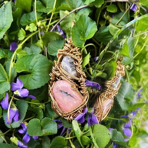Op de afbeelding: Twee edelsteenhangers, elk omhuld met goudkleurig draad. Eén heeft een roze steen, de andere een bruine steen. Ze liggen tegen een achtergrond van groene bladeren en paarse bloemen.