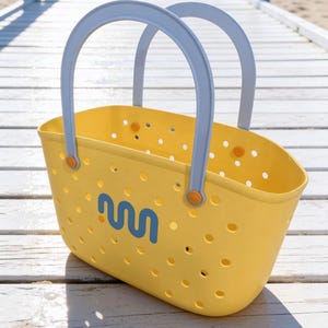 May include: A yellow tote bag with light blue handles and a blue wave-like logo. The bag has small circular perforations and orange accents. The bag is sitting on a white wooden surface.