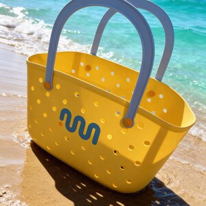 May include: A yellow tote bag with a blue and white logo, gray handles, and small holes. The bag is on a sandy beach with the ocean in the background. The handles are gray and attached with orange fasteners.