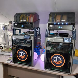 May include: Two grey Creality 3D printers with orange and blue logos, stacked with filament storage. The printers have clear doors and digital displays. Various coloured filament spools are visible, along with other 3D printing equipment on a desk.