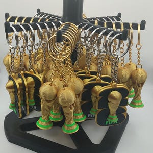 May include: A display of gold-colored trophy keychains with green bases and black accents. The keychains are attached to silver and gold keyrings and chains. The display stand is black.