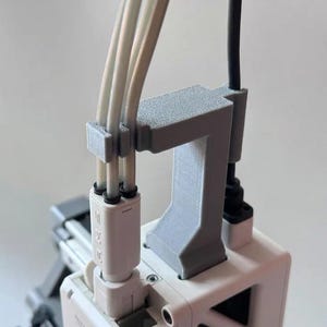 May include: A grey 3D-printed cable organiser holding three white cables and a black cable. The organiser is attached to a white power adapter. The cables are neatly arranged.