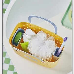 May include: A yellow rectangular caddy with a blue handle, holding toiletries. The caddy contains a green bar of soap, a white pump bottle, a loofah, a tube of toothpaste, and a toothbrush. The caddy is on a white surface.