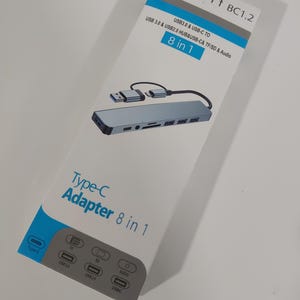 May include: A white rectangular product box for a Type-C Adapter 8 in 1. The box features a graphic of a silver multi-port hub with a short cable. Text includes Support BC1.2, USB 3.0 & USB-C TO USB 3.0 & USB 2.0 HUB & USB-C & TF/SD & Audio.