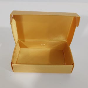 May include: Open, gold-colored box with a satin-like finish. The box is rectangular with a hinged lid, revealing a matching interior. The box is empty and sits on a white surface. The box is likely for jewelry or small gifts.