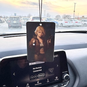 May include: Rectangular car air freshener with a black string, featuring an anime-style illustration of a woman holding a gun. The background is a dark, moody scene. The car's dashboard and a parking lot are visible.
