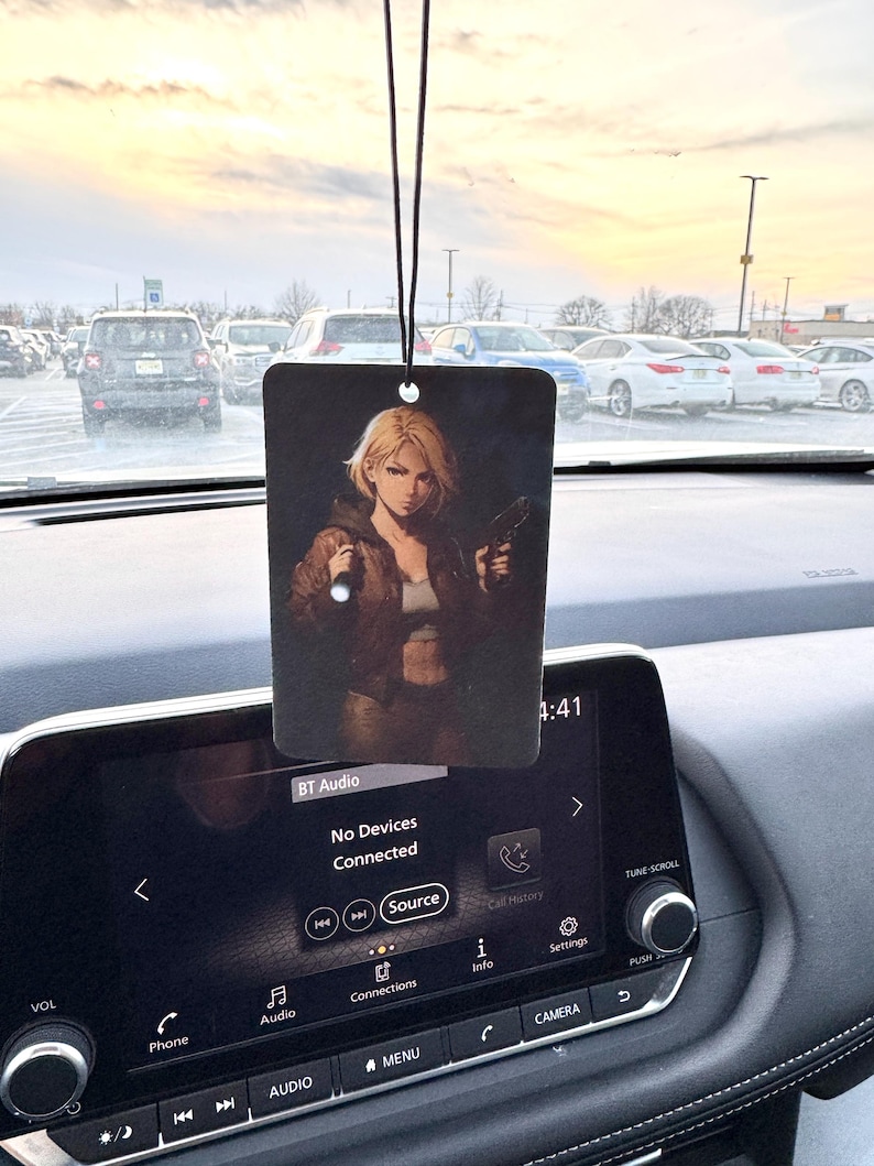 May include: Rectangular car air freshener with a black string, featuring an anime-style illustration of a woman holding a gun. The background is a dark, moody scene. The car's dashboard and a parking lot are visible.