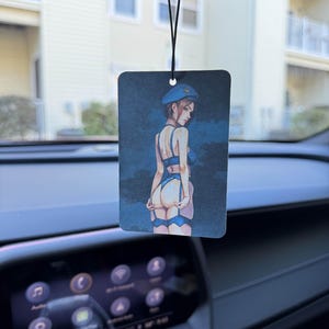 May include: Rectangular car air freshener with a blue and black illustration of a woman in lingerie and a beret. The air freshener hangs from a black string, suspended from a car's rearview mirror.