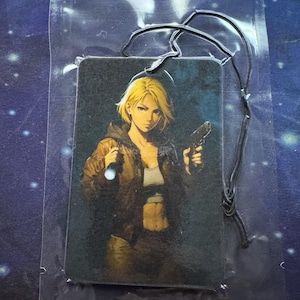 May include: Rectangular air freshener featuring a blonde woman with a gun and a flashlight. The woman is wearing a brown jacket and a white top. The air freshener is in a clear plastic package with a black string.