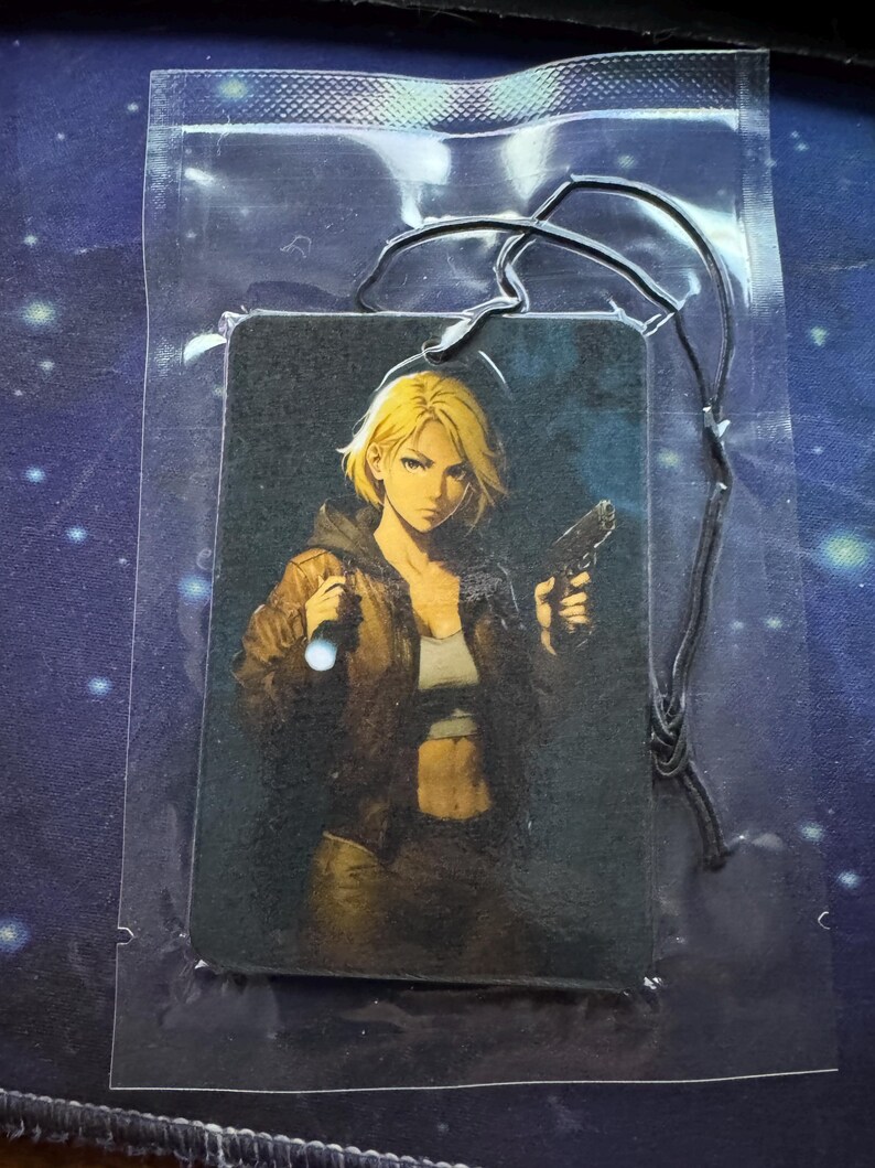 May include: Rectangular air freshener featuring a blonde woman with a gun and a flashlight. The woman is wearing a brown jacket and a white top. The air freshener is in a clear plastic package with a black string.