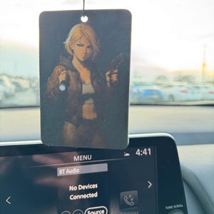 May include: Rectangular air freshener with a printed image of a blonde woman holding a gun. The woman is wearing a brown jacket and a white top. The air freshener is hanging from a black string.