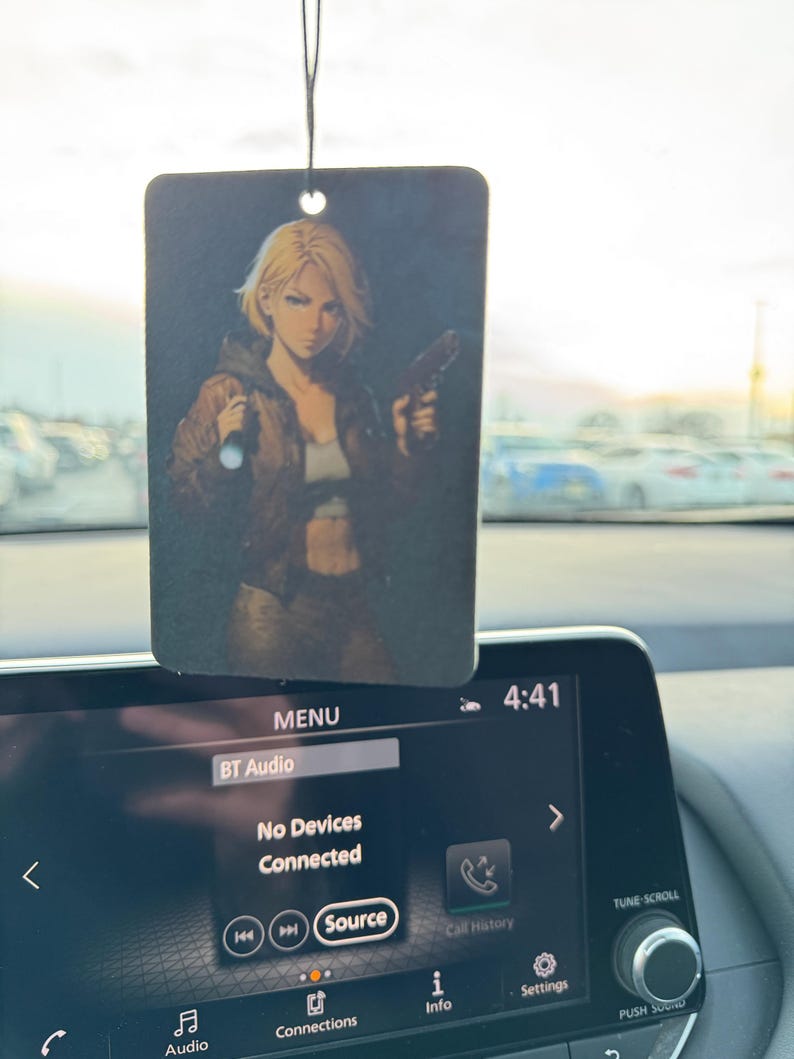 May include: Rectangular air freshener with a printed image of a blonde woman holding a gun. The woman is wearing a brown jacket and a white top. The air freshener is hanging from a black string.