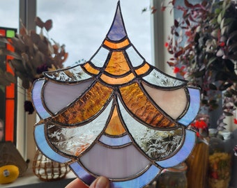 May include: A stained glass suncatcher with a geometric design in shades of purple, orange, and blue. The suncatcher is made of glass and has a metallic finish.