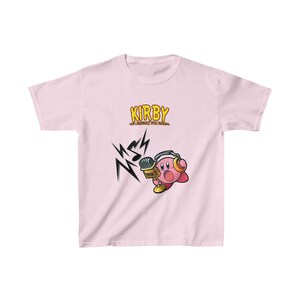 May include: Pink t-shirt featuring a cartoon image of Kirby, a pink character from the video game series, holding a microphone and wearing headphones. The text "KIRBY" and "THE LEGENDARY PINK HERO" is printed in yellow on the shirt.
