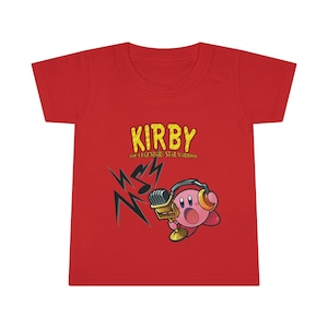 May include: Red t-shirt featuring a cartoon image of Kirby, a pink video game character, singing into a microphone with headphones on. The text "KIRBY THE LEGENDARY STAR WARRIOR" is printed in yellow on the shirt.