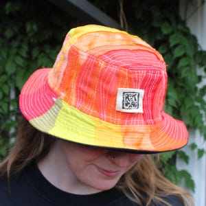 May include: A colorful bucket hat with a vibrant gradient of yellow, orange, and pink hues. The hat features a small square label with text. The brim is lined with black fabric. The hat is a stylish accessory.