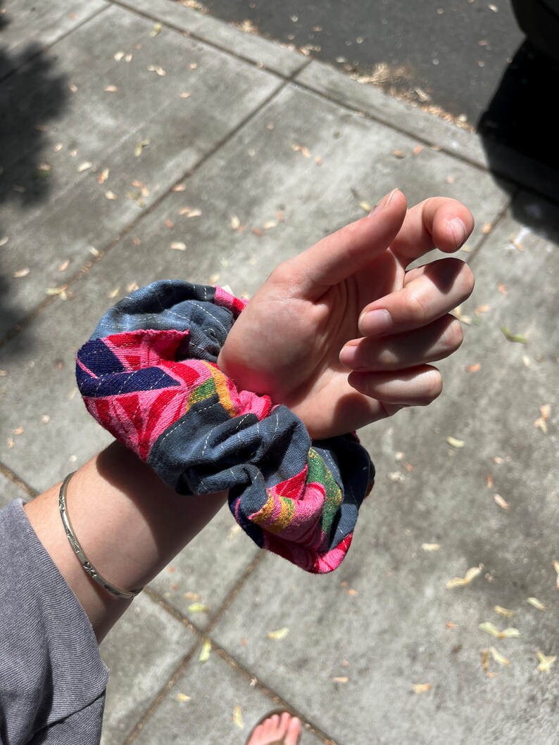 K&ouml;nnte beinhalten: Ein farbenfrohes Haargummi mit einem geometrischen Muster in Rosa, Blau und Gr&uuml;n. Das Haargummi besteht aus Stoff und ist um ein Handgelenk gewickelt. Ein silbernes Armband ist ebenfalls sichtbar.