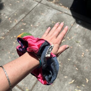 K&ouml;nnte beinhalten: Ein farbenfrohes Haargummi mit einem rosa, blauen und Regenbogenmuster. Das Haargummi ist aus Stoff und hat ein angebrachtes Etikett. Ein silbernes Armband ist ebenfalls sichtbar.