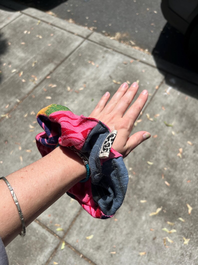 K&ouml;nnte beinhalten: Ein farbenfrohes Haargummi mit einem rosa, blauen und Regenbogenmuster. Das Haargummi ist aus Stoff und hat ein angebrachtes Etikett. Ein silbernes Armband ist ebenfalls sichtbar.