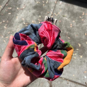 Puede incluir: Una colorida scrunchie con un estampado floral en rosa, rojo, azul y amarillo. La scrunchie se sostiene en una mano y tiene una pequeña etiqueta con un diseño en blanco y negro. Este accesorio para el cabello de tela es un complemento colorido.
