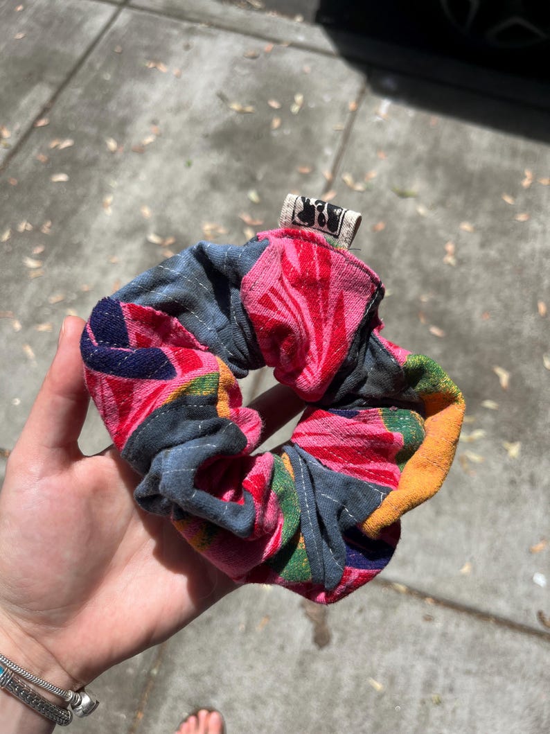 K&ouml;nnte beinhalten: Ein farbenfrohes Haargummi mit einem Blumenmuster in Rosa, Rot, Blau und Gelb. Das Haargummi wird in einer Hand gehalten und hat ein kleines Etikett mit einem Schwarz-Wei&szlig;-Design. Dieses Haaraccessoire aus Stoff ist ein farbenfroher Hingucker.