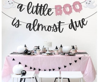 Może przedstawiać: Aranżacja baby shower w różowo-czarnej kolorystyce. Baner głosi "a little BOO is almost due". Stół nakryty jest różowym obrusem, czarno-białymi dekoracjami i białymi stołkami. Różowe balony i dekoracja w kształcie plastra miodu leżą na podłodze.