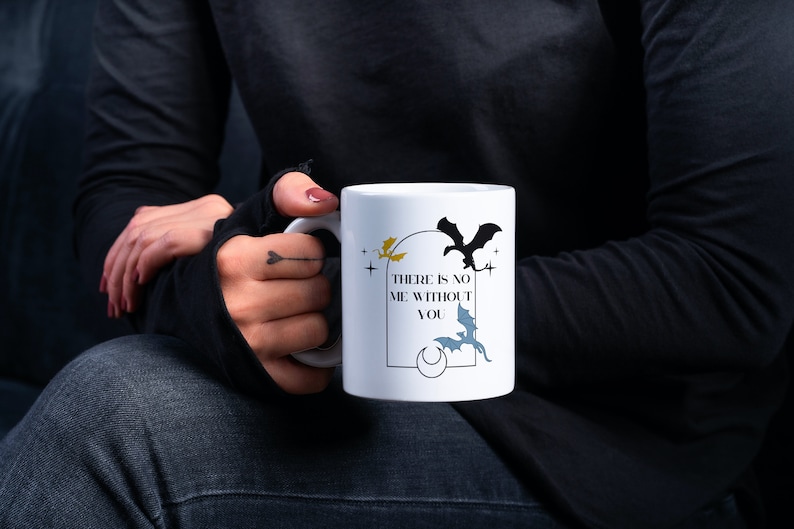 May include: White ceramic mug featuring the text "THERE IS NO ME WITHOUT YOU" and bat illustrations in black, yellow, and blue. The mug is held by a person wearing a black long-sleeve shirt and dark jeans.