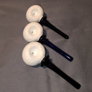 May include: Three white glass smoking pipes with different colored stems. The pipes have a round bowl with a textured surface and a small hole in the center. The stems are black, blue, and dark blue.