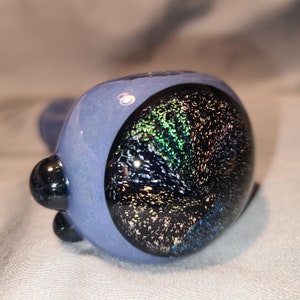 May include: A blue glass pipe with a galaxy-like design. The bowl features a dark, speckled pattern with iridescent colors. The pipe has a rounded bowl and a stem, with a small, black, rounded accent.