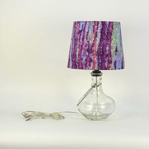 May include: A table lamp featuring a clear glass bottle base and a tapered drum lampshade. The lampshade displays a vertical, abstract pattern in shades of purple, blue, pink, and white. A thin black wire is wrapped around the bottle neck.