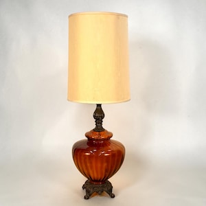 May include: A table lamp featuring a tall, cylindrical beige fabric shade atop a rounded, amber-colored glass base with vertical ribbing. The base is supported by a dark, ornate metal stand with decorative feet and a matching metal neck piece.