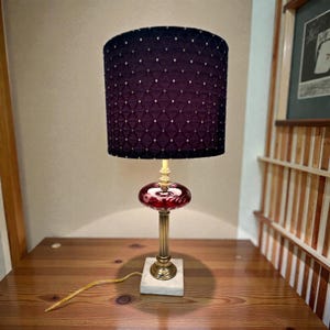 May include: A decorative table lamp with a dark purple lampshade featuring a diamond pattern with small gold dots. The lamp has a red glass center, a brass stem, and a square marble base. The lamp is on a wooden surface.