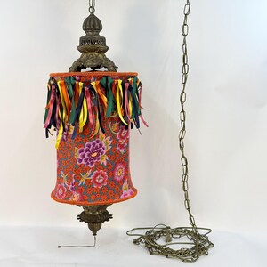 May include: A vintage hanging lamp with a floral patterned shade in red, pink, and purple. The shade is trimmed with colorful ribbons and a brass-colored metal frame. A chain and wire extend from the lamp.