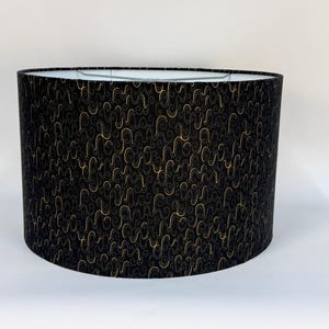 May include: A black oval lampshade with a repeating pattern of gold and gray swirls. The lampshade has a white interior and is designed to fit over a lamp base. The overall shape is cylindrical.