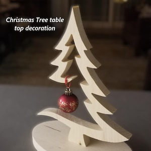 Puede incluir: Una decoración de mesa de madera clara con forma de árbol de Navidad y silueta recortada. Un pequeño adorno rojo redondo con motivos florales dorados cuelga de una rama mediante un hilo rojo. El texto Christmas Tree table top decoration es visible.