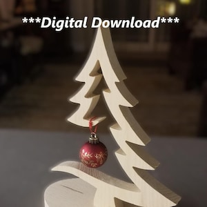 May include: A wooden Christmas tree table top decoration. The tree is cut out of wood and has a red ornament hanging from it. The base of the tree is a round piece of wood.