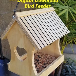 Op de afbeelding: Een houten vogelvoederhuisje met een hartvormige opening en een gestreept dak. Het voederhuisje is gevuld met vogelzaad en hangt aan een ketting. De tekst "*** Digitale Download *** Vogelvoederhuisje -9 bestanden in een Zip-bestand- EPS, AI, SVG, PDF, DXF, CRV" staat onder het voederhuisje.