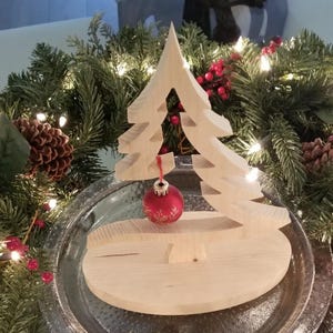 May include: A light-colored wooden Christmas tree cutout standing on an oval wooden base. A small, round, red ornament with a delicate gold-colored floral pattern hangs from the center of the tree by a thin red string, creating a festive display.