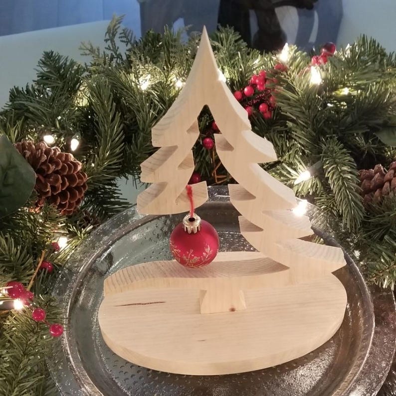 May include: A light-colored wooden Christmas tree cutout standing on an oval wooden base. A small, round, red ornament with a delicate gold-colored floral pattern hangs from the center of the tree by a thin red string, creating a festive display.