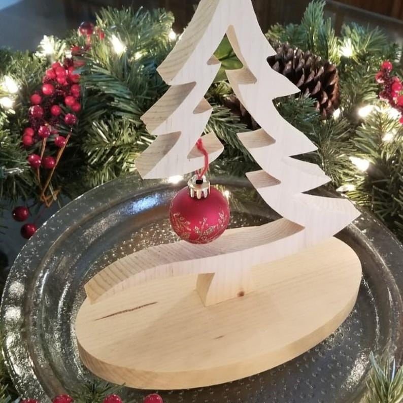 May include: A light-colored wooden Christmas tree cutout standing on an oval wooden base. A single round red ornament with a delicate gold floral pattern hangs from a branch of the tree by a thin red string, centered within the wooden frame.