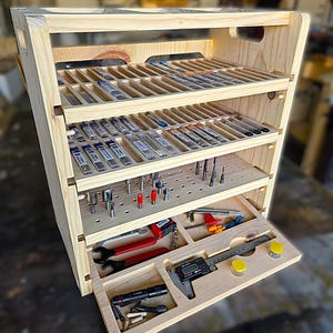 Puede incluir: Un organizador de almacenamiento de madera clara con cuatro niveles. Los dos estantes superiores tienen filas ranuradas para objetos cilíndricos. El tercer estante tiene un diseño de tablero perforado con fresas de metal. El cajón inferior contiene llaves, un calibrador digital, una linterna y herrajes.