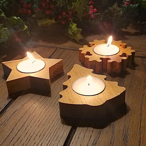 May include: Three wooden candle holders in the shapes of a star, a tree, and a snowflake. Each holder has a small, round opening for a tea light candle. The candles are lit and glowing.