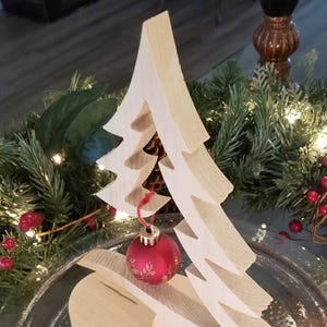 May include: A light-colored wooden Christmas tree cutout with a smooth finish, featuring a single red spherical ornament hanging from a branch by a thin red string. The ornament is decorated with a delicate gold-toned floral pattern.